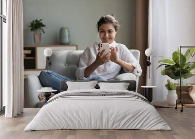 Happy pretty millennial Indian girl relaxing at home, resting in armchair, typing on smartphone, using online app, software, shopping on Internet, making video call. Mobile phone communication concept Wall mural