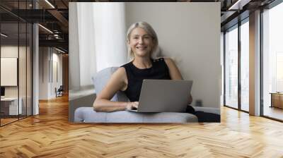 Happy successful mature freelance business woman working from home, holding laptop, looking at camera, smiling, resting in armchair, using modern technology, wireless connection for remote job Wall mural