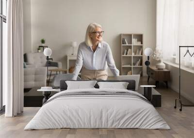 Happy successful senior freelance business woman standing at home workplace table with laptop, looking away with toothy smile, enjoying success, planning, thinking on project strategy, smiling Wall mural