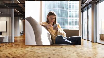 Happy thoughtful millennial gen Z girl using mobile phone at home, sitting on home couch with big window, natural day light in background, enjoying shopping, online communication Wall mural