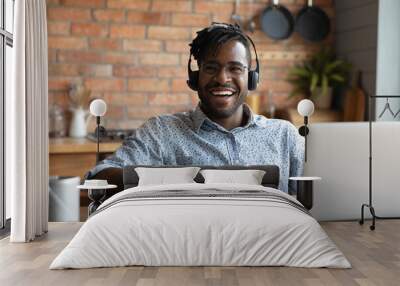 Happy young african american man in eyewear and modern headset sitting at table with computer, laughing distracted from distant study or remote job, having fun entertaining feeling joyful at home. Wall mural