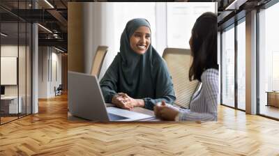 Happy young Arab business professional woman in hijab talking to European female colleague at meeting table, smiling. Two young diverse businesswomen discussing teamwork, work project Wall mural