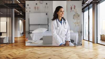 Happy young cardiologist woman posing at workplace, leaning on clinic office table, looking away, smiling, enjoying occupation, thinking on successful doctor career. Casual portrait Wall mural