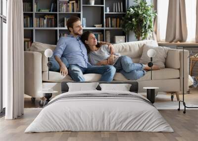 Happy young Caucasian couple renters or tenants relax on couch in living room look in distance dreaming thinking. Smiling dreamy man and woman rest on sofa at home imagine visualize together. Wall mural