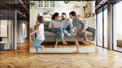 Happy young couple sitting at couch near running children using laptop in modern living room. Brother and sister playing in catch up near mother and father at home. Family having fun together. Wall mural