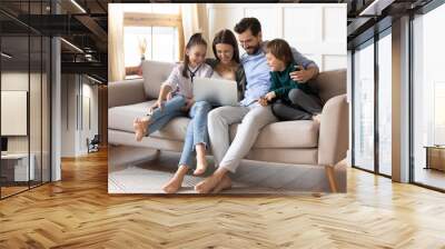 Happy young family with two small kids relax on sofa in living room at home have fun using laptop together. Smiling Caucasian parents with little children talk speak on webcam video call on computer. Wall mural