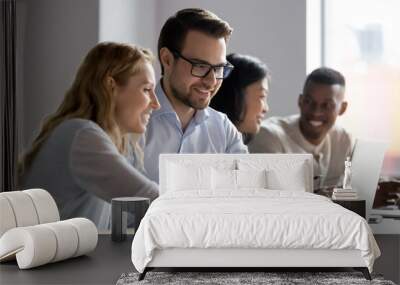 Happy young female employee discussing online project, showing computer presentation to skilled team leader in eyeglasses. Friendly diverse colleagues working in pairs on laptop, using applications. Wall mural