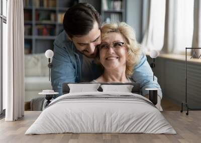 Happy young man hugging mature mother wearing glasses from back, expressing love and gratitude, adult son and middle aged woman cuddling, family enjoying tender moment, sitting in living room at home Wall mural