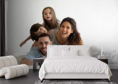 Happy young parents with little children lying on bed looking at camera. Smiling beautiful mother and bearded father enjoying free time with cute adorable daughter and son. Wall mural