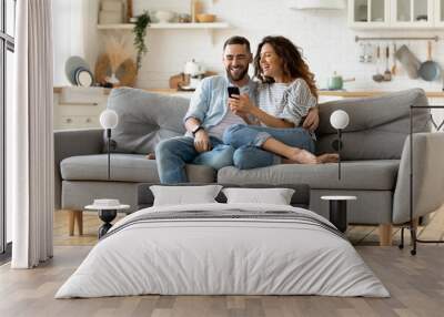 Happy young woman and man hugging, using smartphone together, sitting on cozy couch at home, smiling overjoyed wife and husband looking at phone screen, sitting on sofa in modern living room Wall mural