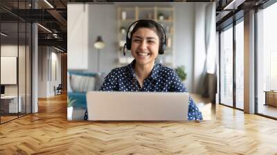 Head shot cheerful smiling pretty hindu ethnic girl sitting at table with computer, wearing headphones with mic, looking at camera. Happy indian woman professional tutor educating client online. Wall mural