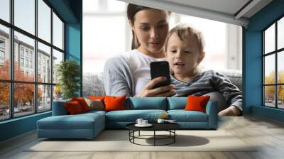 Head shot pleasant young woman cuddling little preschool kid son, taking selfie on telephone. Smiling mommy nanny babysitter showing cartoons or mobile apps to small child boy, resting in living room. Wall mural