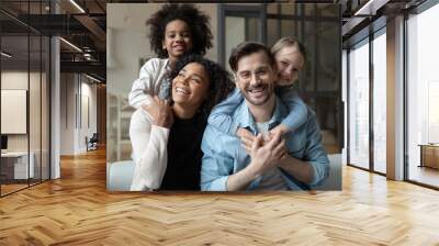 Head shot portrait happy multiracial family looking at camera, African American wife and Caucasian husband piggy backing two little daughters, posing for photo, having fun, sitting on couch Wall mural