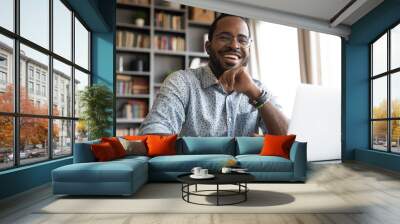 Head shot portrait laughing African American businessman wearing glasses sitting at work desk with laptop, looking at camera, handsome happy young man freelancer blogger working online Wall mural