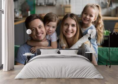 Head shot portrait of happy parents piggybacking son and daughter sitting on sofa together, smiling family with cute kids bonding embracing at home, children hugging mother father looking at camera Wall mural