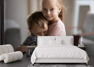 Head shot portrait of little kid girl cuddling smaller brother at home, showing love and care. Compassionate sister comforting soothing upset stressed boy in living room, siblings relations concept. Wall mural