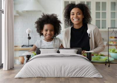 Head shot portrait smiling African American mother with adorable little daughter cooking salad together, chopping vegetables, family sitting at wooden table in kitchen, looking at camera Wall mural