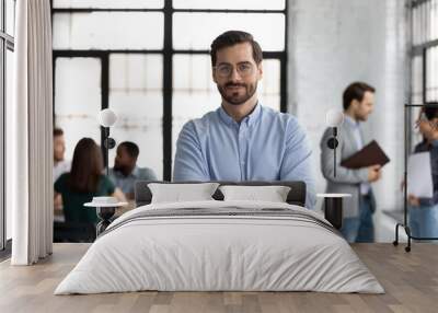 Head shot portrait smiling confident businessman wearing glasses standing in modern office room with arms crossed, diverse colleagues on background, executive boss startup founder looking at camera Wall mural