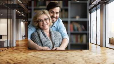 Head shot portrait smiling mature woman and young man hugging, looking at camera, loving adult son embracing elderly mother from back, standing in modern living room together, posing for photo Wall mural