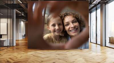 Head shot portrait smiling mother and little daughter taking selfie together, looking through fingers at camera, making heart shape gesture, happy mom and adorable girl kid bonding, expressing love Wall mural