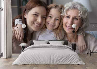 Head shot portrait smiling pretty little girl hugging young mother and mature grandmother, three generations of women looking at camera, posing for family photo, sitting on couch at home Wall mural