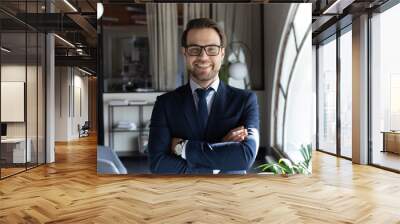 Head shot smiling confident young businessman executive wearing glasses and suit standing in modern office room, happy successful entrepreneur employee intern with arms crossed looking at camera Wall mural