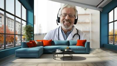 Headshot portrait of smiling mature Caucasian male doctor in headphones talk on video call in hospital. Profile picture of happy senior man GP or therapist in earphones consult patient on webcam. Wall mural