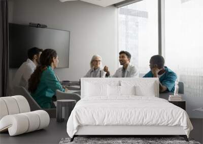 Indian male therapist wear white coat makes speech at group meeting medical seminar in modern conference room. Multiethnic clinic staff members listening opinion, decisions and solutions of workmate Wall mural