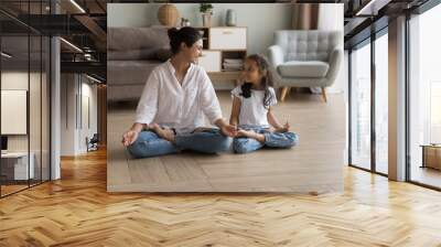 Indian woman laughing seated cross-legged on floor doing meditation practice with cute little daughter at modern cozy home. Loving parent teach child yoga, instill good habits for healthy lifestyle Wall mural