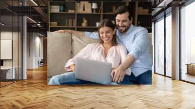 Joyful happy married couple relaxing at laptop on couch, watching funny movie, browsing internet, shopping online, using ecommerce app, making video call, enjoying leisure together, laughing, smiling Wall mural