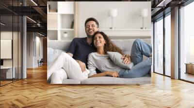 Joyful married couple in love resting on comfortable sofa, hugging with affection, trust, talking, laughing, having fun together, enjoying romantic leisure, boning, romance at home Wall mural