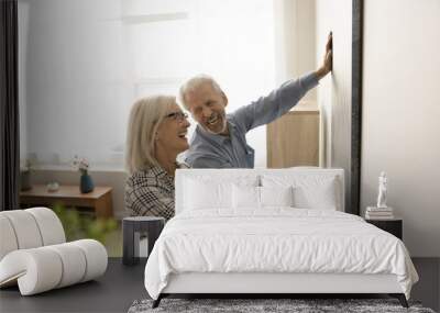 Joyful positive retired husband and wife enjoying interior decoration work, hanging large artwork on wall, holding picture together, smiling, laughing, having in new apartment Wall mural