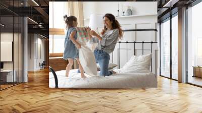 Laughing young mother and little daughter playing pillow fight, jumping on bed in bedroom, cute preschool girl and happy young mum having fun together, funny family activity on weekend Wall mural