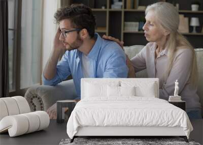 Loving older woman comforting her upset young adult son, gives wise advice in difficult life situation, talking, provide psychological support showing care seated on couch, spend time together at home Wall mural