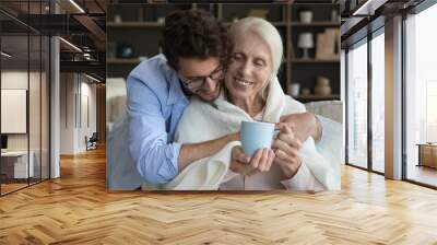 Loving young adult son giving positive sick senior mother help, assist, care, wrapping mom shoulders into plaid, scarf, giving cup of hot drink, hugging, smiling, laughing Wall mural