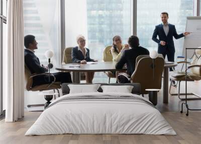 Male employee give flipchart presentation to smiling colleagues at negotiations, man present project or idea to happy coworkers at briefing, workers laugh at office meeting, having casual talk Wall mural