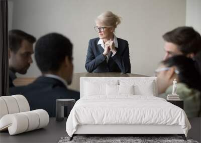 Middle aged stressed woman sitting at the desk in office at meeting. Young multiracial workers team sitting their backs to camera. Passing interview, hiring, recruiting and age discrimination concept Wall mural