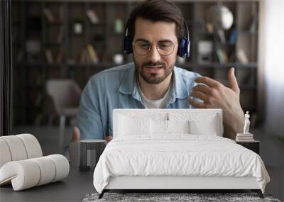 Millennial Caucasian man in earphones look at laptop screen talk on video call on gadget. Young male in headphones have webcam digital virtual online conference on computer. Technology concept. Wall mural