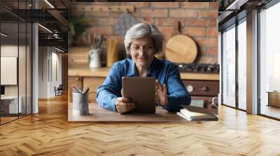 Modern day pensioner. Smiling mature latina woman sit at home office by kitchen table watch video on tab computer screen. Old age grandmother enjoy reading e book on tablet pc scroll news on websites Wall mural