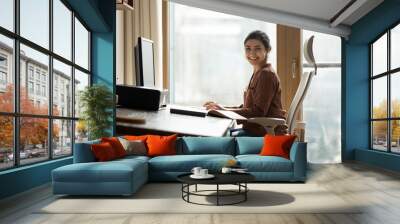 Modern day worker. Portrait of happy biracial business woman freelancer sit by computer at comfy workplace at corporate workspace or at home. Smiling young indian lady office employee look at camera Wall mural