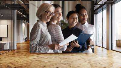 Motivated multiracial female team office workers of different generations look at one side smile ready to meet help client. Professional diverse businesswomen stand together hold documents clipboards Wall mural