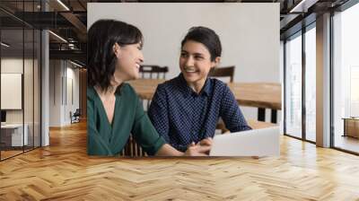 Multi ethnic women colleagues laughing in office at workplace desk, share thoughts, ideas, opinions enjoy break and pleasant informal talk during workday, having successful teamwork, enjoy friendship Wall mural