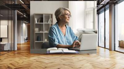 Older woman sits at desk using her laptop, engaged in digital activities, looking thoughtfully out window, studying, working, or managing personal tasks online in cozy domestic room. Tech, lifestyle Wall mural