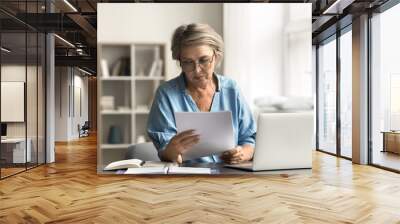 Older woman with serious expression read document sitting at desk with laptop, dealing with important paperwork, managing finances or health-related issues, learn written information looks concerned Wall mural