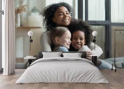 Overjoyed African American woman cuddling with two little daughters, multiracial family enjoying tender moment, sitting on cozy couch at home, loving mum hugging kids, adopted child Wall mural