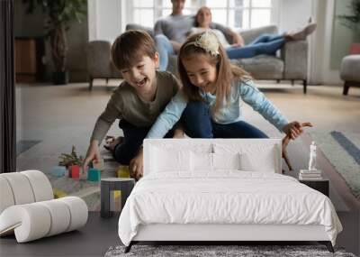 Overjoyed funny little children siblings playing toys on floor carpet while parents resting on sofa. Happy emotional cute little boy having fun with preschool sister, enjoying playtime in living room. Wall mural