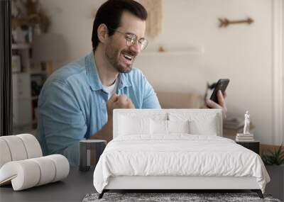 Overjoyed man wearing glasses holding smartphone, showing yes gesture, celebrating success close up, business achievement, lottery win, looking at phone screen, reading great news in email or message Wall mural