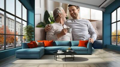 Overjoyed mature father and adult son sit relax on couch at home drink coffee or tea talking on weekend. Happy elderly dad and grownup man child have fun laugh enjoying family time together. Wall mural