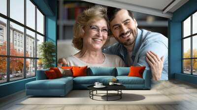 Overjoyed mature woman wearing glasses and adult son hugging, posing for photo, hugging, sitting on couch in living room, excited happy middle aged mother and young man embracing, two generations Wall mural