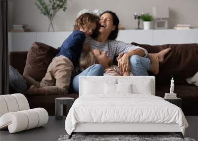 Overjoyed mother with two kids having fun on comfortable couch at home, laughing happy mom with adorable daughter and son tickling, engaged in funny active game, spending leisure time together Wall mural